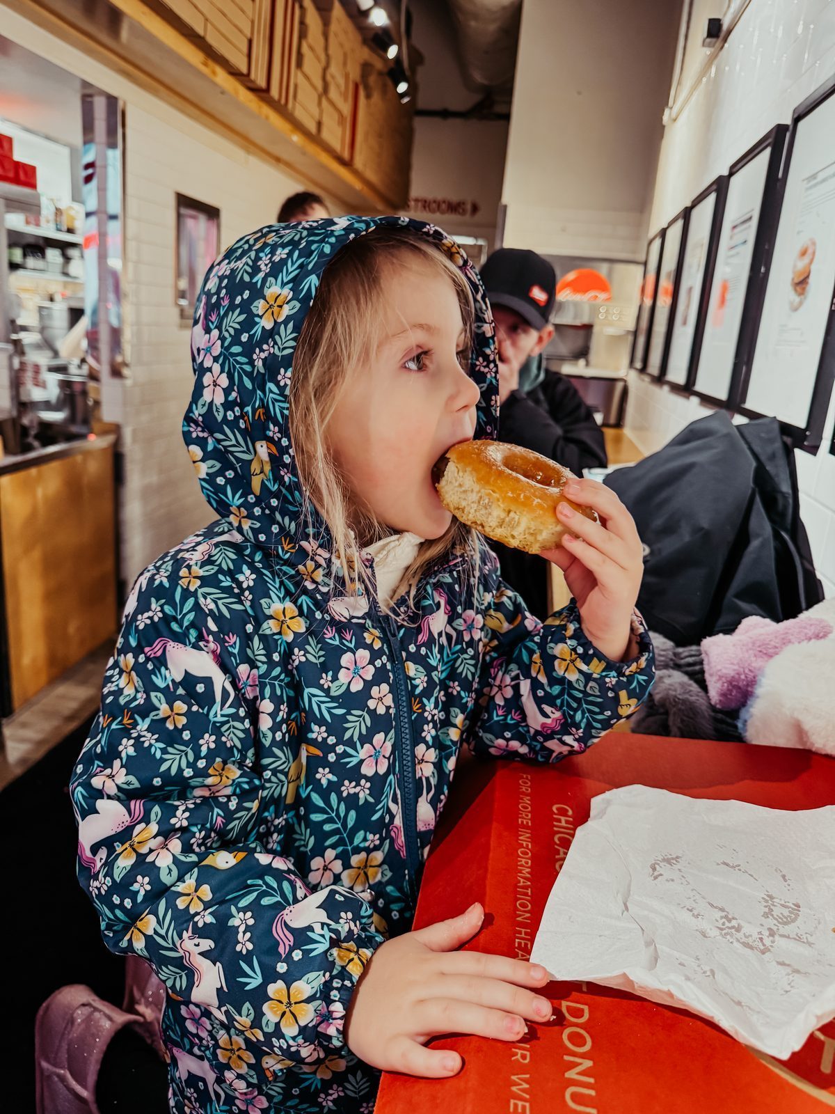 A girl in a unicorn coat taking a bite of her donut